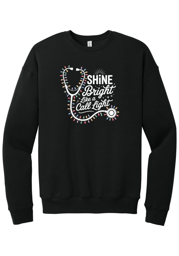 Shine Bright Like a Call Light crewneck sweatshirt in Black, featuring a white stethoscope wrapped in multicolor Christmas lights with Shine Bright Like a Call Light lettering and sparkles