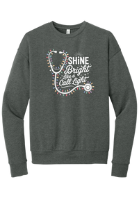 Shine Bright Like a Call Light crewneck sweatshirt in Deep Heather, featuring a white stethoscope wrapped in multicolor Christmas lights with Shine Bright Like a Call Light lettering and sparkles
