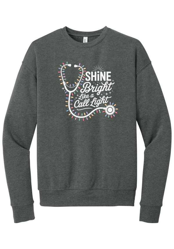 Shine Bright Like a Call Light crewneck sweatshirt in Deep Heather, featuring a white stethoscope wrapped in multicolor Christmas lights with Shine Bright Like a Call Light lettering and sparkles