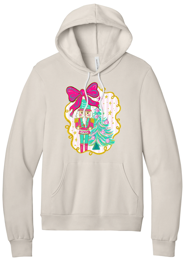 Nutcracker Bow hoodie in Heather Dust, featuring a teal and pink nutcracker beside a tinsel tree under a big hot pink bow