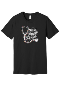 Shine Bright Like a Call Light tee in Black, featuring a white stethoscope wrapped in multicolor Christmas lights with bold Shine Bright Like a Call Light lettering