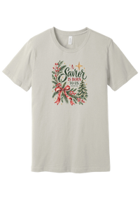 A Savior Is Born To Us tee in Dust, featuring watercolor evergreen, holly, and a red ribbon bow with hand-lettered A Savior is born to us and Isaiah 9:6