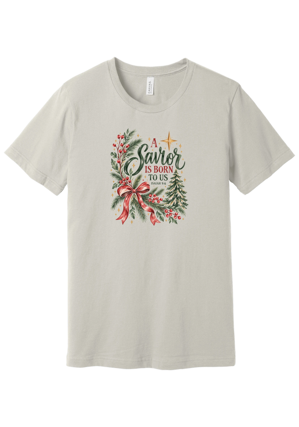 A Savior Is Born To Us tee in Dust, featuring watercolor evergreen, holly, and a red ribbon bow with hand-lettered A Savior is born to us and Isaiah 9:6